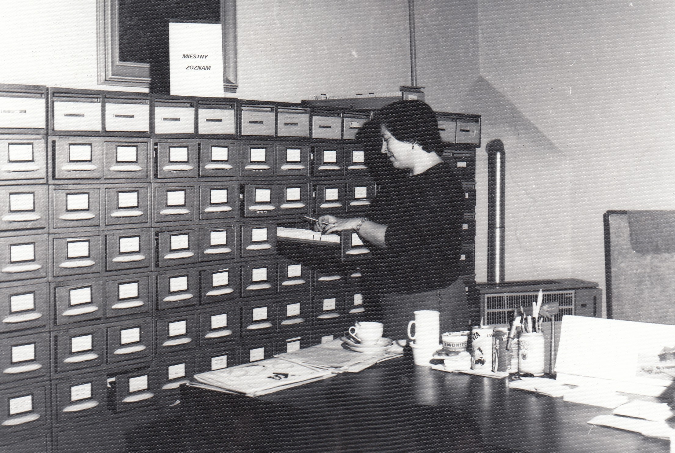 Knihovníčka na fotke zo sedemdesiatych rokov prezerá kartotečné lístky v kartotečnom regáli, ktorý zaberá celú fotku.